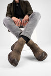 Person wearing brown leather boots, gray jeans, and a dark jacket on a white background